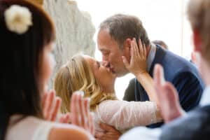 Ein Paar küsst sich freudig bei seiner Hochzeitszeremonie. Die Frau mit blonden Haaren und Spitzenkleid küsst den Mann im Anzug, während sie von applaudierenden Gästen umgeben sind.
