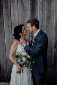 Braut und Bräutigam stehen eng beieinander vor einer Holzwand und verkörpern so die Essenz ihrer Hochzeit. Die Braut hält einen Strauß weißer Blumen und Grünpflanzen in der Hand, während sie sich in die Augen blicken und innig lächeln. Der Bräutigam trägt einen dunklen Anzug, während die Braut ein atemberaubendes weißes Kleid trägt.