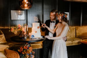 Braut und Bräutigam schneiden glücklich ihre Hochzeitstorte an, ein perfekter Moment ihrer Hochzeit. Die Braut trägt einen Blumenkranz und ein weißes Kleid, während der Bräutigam einen Anzug trägt. Die Umgebung ist warm beleuchtet mit Blumen und Kerzen auf dem Tisch neben der Torte, alles Teil einer sorgfältigen Hochzeitsplanung.