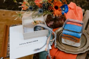 Ein farbenfrohes Arrangement für eine Hochzeit besteht aus einem farbenfrohen Blumenstrauß, einem Buch mit dem Titel „Nina Walcher: Ursprung Reden“, einer kleinen offenen Holzkiste, gemusterten Stoffbändern und einem Glasgefäß, das mit blauem und orangefarbenem Sand bedeckt ist. Perfekt für alle, die sich mit sorgfältiger Hochzeitsplanung beschäftigen.
