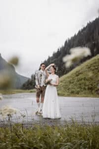 Ein Paar steht auf einer Bergstraße, umgeben von Grün, und feiert seine Hochzeit. Der Mann in traditioneller Kleidung hält liebevoll die Hand der Frau, die mit einem Blumenstrauß lächelt. Es ist ein freudiger Moment in dieser ruhigen, natürlichen Umgebung.