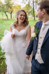 Braut und Bräutigam gehen mitten in ihrer Hochzeit Hand in Hand einen von Bäumen gesäumten Weg entlang. Die Braut strahlt in einem weißen Kleid und lächelt neben ihrem Bräutigam im marineblauen Anzug mit weißer Weste und Ansteckblume. Saftiges grünes Gras und Bäume bilden die perfekte Kulisse für ihren besonderen Tag.
