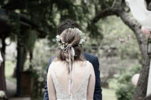 Eine Braut mit floralem Haarschmuck steht ihrem Bräutigam im blauen Anzug im Freien gegenüber. Die Szene, perfekt für jeden Hochzeitsplanungsmoment, ist von Bäumen und Grün umgeben und schafft eine ruhige, natürliche Kulisse.