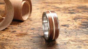 Ein Silberring mit Holzeinlage liegt auf einer rustikalen Holzoberfläche, perfekt für alle, die ihre Hochzeit planen. Im Hintergrund befindet sich ein gerolltes Stück Holzfurnier, das eine handwerkliche Note verleiht.