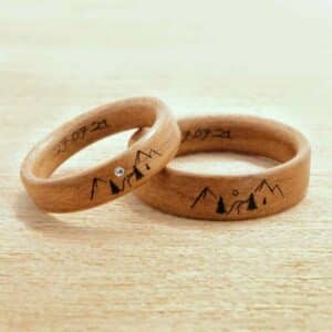 Zwei Holzringe liegen auf einer hellen Holzoberfläche, perfekte Symbole für Ihre Hochzeit. Jeder Ring ist mit Berg-, Baum- und Sonnengravuren versehen, während einer einen kleinen Edelstein und das Datum „21-07-21“ enthält, das innen eingraviert ist. Ideal für Ihre Hochzeitsplanung.