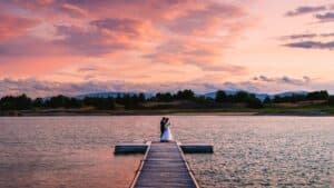 Ein Paar steht am Ende eines Holzstegs an einem See und umarmt sich unter einem leuchtend rosa und orangefarbenen Sonnenuntergangshimmel – eine perfekte Szene für ihre Hochzeit. Das ruhige Wasser spiegelt die bunten Wolken und die umgebenden Silhouetten der entfernten Bäume und Hügel wider und fängt die Romantik von Heiraten ein.