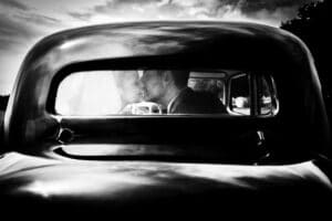 Ein Schwarzweißfoto eines frischvermählten Paares, das sich in einem Oldtimer küsst, fängt die Essenz der Romantik einer Hochzeit ein. Die Aufnahme aus der Heckscheibe fängt ihren zärtlichen Moment vor dem Hintergrund eines bewölkten Himmels ein und verkörpert die zeitlose Schönheit von Heiraten.