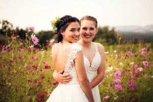 Zwei Frauen in Brautkleidern umarmen sich und lächeln auf einer farbenprächtigen Blumenwiese und verkörpern so die Freude ihrer Hochzeit. Der Hintergrund zeigt eine malerische Landschaft unter einem klaren Himmel, perfekt für ihren besonderen Tag.
