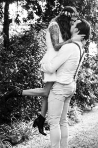 Auf einem zeitlosen Schwarzweißfoto, das die Essenz des Hochzeitsglücks einfängt, küsst sich ein Paar inmitten üppigen Grüns. Die Frau, geschmückt mit einem kurzen Kleid und einer Blumenkrone, wird von ihrem Partner in Hosenträgern hochgehoben und verkörpert die Freude des Heiratens in einer idyllischen Umgebung.