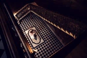 Braut und Bräutigam umarmen sich auf einem schwarz-weiß karierten Boden, umgeben von einer großen Treppe mit aufwendigem Geländer. Die Szene verkörpert die Schönheit sorgfältiger Hochzeitsplanung, aufgenommen von oben in einer schwach beleuchteten, eleganten Umgebung.