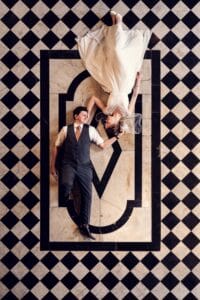 Eine Braut im weißen Kleid und ein Bräutigam in Weste und Krawatte liegen auf einem schwarz-weiß karierten Boden, ihre Köpfe berühren sich romantisch. Diese perfekte Komposition fängt die Essenz der Hochzeitseleganz ein, ein inspirierender Moment für jedes Paar, das mitten in der Hochzeitsplanung steckt.