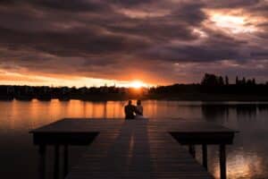 Ein Paar sitzt am Ende eines Holzstegs und bespricht vielleicht seine Hochzeitsplanung, während sie einen dramatischen Sonnenuntergang über einem ruhigen See beobachten. Der Himmel ist voller dunkler Wolken und leuchtender Orangetöne, die sich auf der Wasseroberfläche spiegeln. In der Ferne säumen die Silhouetten von Bäumen den Horizont.