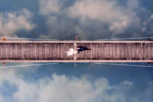 Luftaufnahme eines Paares, das auf einer Holzbrücke über einem ruhigen Gewässer liegt und von seiner Hochzeit träumt. Händchenhaltend, unter ihnen spiegeln sich weiche Wolken, finden sie Gelassenheit in der magischen Atmosphäre, die perfekt für eine Hochzeit ist.