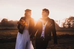 Während sie zur goldenen Stunde Hand in Hand durch ein Feld spazieren, verkörpert das Paar Romantik. Die Frau in einem weißen Kleid mit Sonnenblumen in der Hand und ihr Partner tragen beide schwarze Jacken. Die Sonne scheint warm hinter ihnen und fängt einen idyllischen Moment ein, der perfekt für jede Hochzeitsplanung ist.