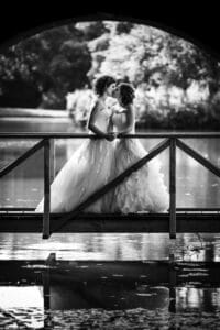 Zwei Frauen in Brautkleidern stehen auf einer Brücke und umarmen sich innig, während das Sonnenlicht durch die Bäume fällt und eine romantische Atmosphäre schafft. Während sie über ihren Weg von der Hochzeitsplanung zur Freude am Heiraten nachdenken, schimmern ihre Spiegelbilder im Wasser unter ihnen.