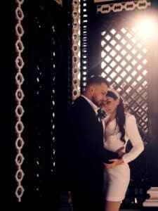 Ein Paar umarmt sich in einer schwach beleuchteten Umgebung mit dekorativen Gittermustern und verkörpert die Essenz einer perfekten Hochzeit. Der Mann trägt einen Anzug, während die Frau ein weißes Kleid trägt. Sonnenlicht fällt hindurch und wirft von hinten einen warmen Schein auf sie.