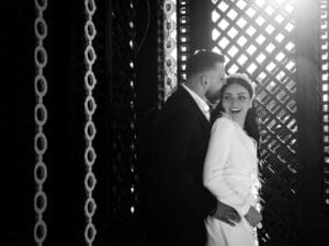 Ein Paar steht dicht beieinander und lächelt fröhlich in einer sonnenbeschienenen Hochzeitskulisse mit komplizierten Gittermustern und runden Lampen im Hintergrund. Die Frau trägt ein weißes Kleid und blickt über ihre Schulter. Die Szene vermittelt ein Gefühl von Glück und Romantik.