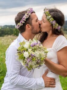 Auf einer üppigen grünen Wiese unter einem bewölkten Himmel küsst sich das Paar zärtlich, ihre Blumenkränze symbolisieren ihre freudige Hochzeit. Der Bräutigam im weißen Hemd umarmt seine Braut, die einen Strauß weißer, violetter und grüner Blumen hält, während sie dieses schöne Kapitel der Hochzeit feiern.