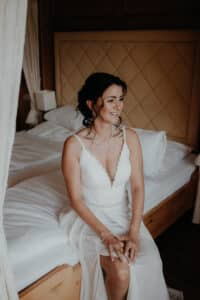 Eine Braut in einem weißen Brautkleid sitzt auf der Bettkante und verkörpert die heitere Eleganz ihres Hochzeitstages. Ihr dunkles Haar ist in lockeren Locken gestylt und wird durch minimalen Schmuck ergänzt. Das Zimmer verfügt über ein gestepptes Kopfteil und transparente Vorhänge, die eine perfekte Atmosphäre für einen so bedeutsamen Anlass schaffen.