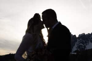 Silhouette eines sich küssenden Paares vor einer Berglandschaft. Der Himmel, sanft erhellt von der aufgehenden oder untergehenden Sonne, wirft ein sanftes Gegenlicht – perfekt, um den Moment des Heiratens festzuhalten. Die Frau trägt einen Blumenkopfschmuck und der Mann ist im Anzug.