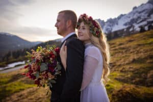 Eine Braut in einem weißen Kleid und einer Blumenkrone umarmt einen Bräutigam im schwarzen Anzug von hinten und verkörpert die Essenz einer Hochzeit vor der Kulisse schneebedeckter Berge. Sie hält einen farbenfrohen Blumenstrauß auf dem grasbedeckten Hügel. Die Szene ist ruhig und malerisch, perfekt für diejenigen, die in die Hochzeitsplanung vertieft sind.