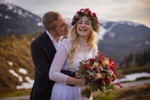 Ein Paar in einer Berglandschaft. Die Frau, geschmückt mit einem Blumenkranz und einem weißen Kleid, lächelt und hält einen Blumenstrauß. Der Mann steht dahinter und umarmt sie in dieser malerischen Umgebung, die perfekt für die Feier ihrer Hochzeit ist, mit Schneeflecken und Kiefern im Hintergrund.
