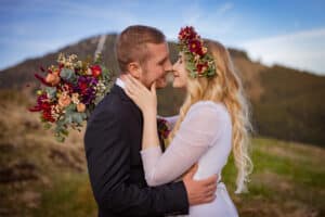 Ein Paar in Hochzeitskleidung steht eng beieinander und lächelt sich während der Hochzeit an. Der Bräutigam hält einen Blumenstrauß und die Braut trägt eine Blumenkrone. Sie sind draußen mit einem Berg und klarem Himmel im Hintergrund und erwecken ihre sorgfältig geplante Hochzeitsplanung zum Leben.
