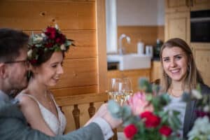 Braut und Bräutigam erheben inmitten ihrer Hochzeitsfeier mit einer lächelnden Frau ihre Champagnergläser. Die Braut trägt einen Blumenkopfschmuck, der die Freude am Heiraten verkörpert. Die Atmosphäre ist warm und gemütlich mit Holzvertäfelung und einer Blumenvase im Vordergrund.