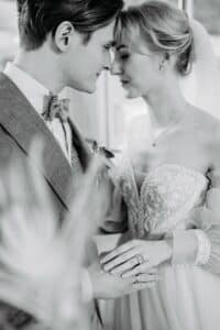 Ein Schwarzweißfoto fängt die Essenz einer Hochzeit ein, bei der Braut und Bräutigam eng beieinander stehen und ihre Stirnen berühren. Die Braut trägt ein Spitzenhochzeitskleid und einen Schleier und hält die Hand des Bräutigams. Der Bräutigam trägt einen Anzug mit Fliege; beide lächeln sich sanft an und verkörpern damit die Freude des Heiratens.