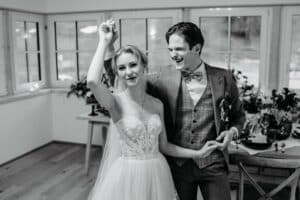 Ein fröhliches Paar tanzt während seines Hochzeitsempfangs in einem elegant dekorierten Raum. Die Braut trägt ein trägerloses Spitzenkleid und der Bräutigam Anzug und Fliege. Sie lächeln und genießen den Moment, umgeben von natürlichem Licht.