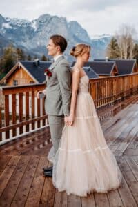 Braut und Bräutigam stehen Rücken an Rücken auf einem Holzdeck und halten einen perfekten Hochzeitsmoment fest. Der Bräutigam trägt einen hellgrauen Anzug und die Braut glänzt in ihrem trägerlosen Kleid, während schneebedeckte Berge und rustikale Hütten eine bezaubernde Kulisse für ihren besonderen Tag bilden.