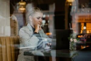 Eine Frau in einem karierten Blazer sitzt an einem Cafétisch, konzentriert auf ihren Laptop und ist wahrscheinlich in die Hochzeitsplanung vertieft. Auf dem Tisch steht ein Glas mit rosa Blumen und einem Strohhalm. Warmes Licht und Reflexionen evozieren eine Atmosphäre, die perfekt zum Träumen von Hochzeitsfeiern ist.