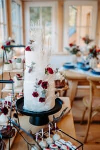 Eine dreistöckige weiße Hochzeitstorte, geschmückt mit tiefroten Blumen und dekorativen Federn, wird elegant auf einem schwarzen Ständer präsentiert. Auf dem Tisch stehen außerdem verschiedene Gebäcke und Desserts, die eine gemütliche Atmosphäre schaffen, die perfekt für eine stilvolle Hochzeit ist.