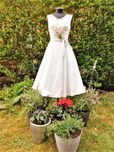 Eine schwarze Schaufensterpuppe präsentiert ein weißes Kleid mit einem Blumenarrangement im Ausschnitt und steht inmitten mehrerer Blumentöpfe in einem Garten – eine perfekte Vision für eine Hochzeit. Der Hintergrund ist eine üppige grüne Hecke, die ideale Inspiration für alle, die an der Hochzeitsplanung beteiligt sind.