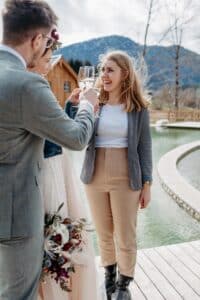 Eine Gruppe von drei Personen steht an einem Pool, stößt mit Weingläsern an und feiert die Freude der Hochzeit. Eine Frau lacht und trägt einen Blazer und hellbraune Hosen. Ein Mann in einem grauen Anzug und eine Frau in einem weißen Kleid und einer Jeansjacke sind teilweise zu sehen. Berge bilden den Hintergrund.