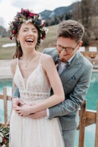 Ein fröhliches Paar feiert seine Hochzeit in der Nähe eines malerischen Außenpools. Die Braut, die einen Blumenkranz und ein Spitzenkleid trägt, lacht, während der Bräutigam mit Brille und leichtem Anzug sie liebevoll von hinten umarmt. Majestätische Berge und Bäume bilden die perfekte Kulisse für diesen besonderen Moment.