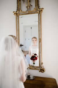 Eine Braut in Brautkleid und Schleier steht vor einem verzierten Spiegel und hält einen Strauß roter und weißer Blumen in der Hand. Ihr Spiegelbild fängt ihren heiteren Gesichtsausdruck ein und unterstreicht die Freude an der Hochzeit, während sie diesen perfekten Moment inmitten einer sorgfältigen Hochzeitsplanung genießt.