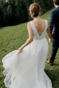 Eine Braut in einem weißen Kleid mit Blumendetails hält die Hand eines Mannes im Anzug und verkörpert damit die Essenz einer Hochzeit. Sie schlendern über eine von Bäumen umgebene Wiese. Ihr Haar ist mit kleinen Blumen geschmückt, während sie mit der anderen Hand elegant einen Teil ihres Kleides anhebt.