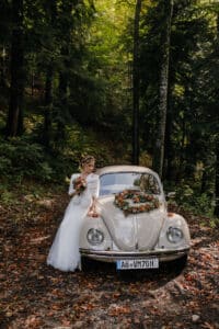 Eine Braut in einem weißen Kleid mit einem Blumenstrauß lehnt an einem Oldtimer, der wunderschön mit Blumen geschmückt ist. Das Auto ist auf einem laubbedeckten Weg geparkt, umgeben von üppigem, grünem Wald – die perfekte Kulisse für einen unvergesslichen Hochzeitsmoment.