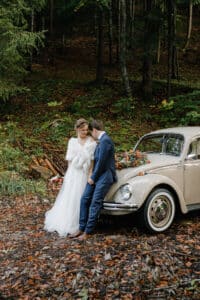 In einer ruhigen Waldlandschaft lehnen die Braut in ihrem weißen Kleid und der Bräutigam in seinem blauen Anzug an einem mit Blumen geschmückten Oldtimer und teilen einen zärtlichen Moment inmitten gefallener Blätter. Diese bezaubernde Szene fängt die Essenz ihrer Hochzeit wunderschön ein.