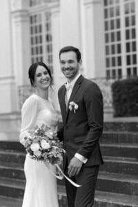 Braut und Bräutigam stehen fröhlich lächelnd auf einer Treppe und feiern ihre Hochzeit. Die Braut trägt ein langes weißes Kleid und hält einen Blumenstrauß, während der Bräutigam einen Anzug mit einer Ansteckblume trägt. Die Kulisse ist geprägt von großen Fenstern und verzierten Metallgeländern. Schwarzweißfoto.