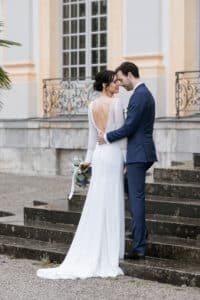 Eine Braut in einem langen weißen Kleid mit offenem Rücken steht mit einem Bräutigam in einem marineblauen Anzug auf einer Treppe im Freien. Als Teil ihrer Hochzeit blicken sie einander an und halten sich an den Händen. Im Hintergrund sind ein großes Gebäude und ein dekoratives Geländer zu sehen. Die Braut hält einen Blumenstrauß.