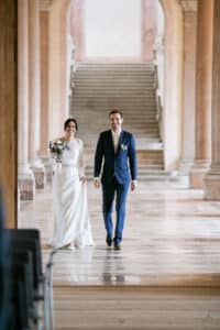 Eine Braut in einem weißen Kleid hält Blumen in der Hand und geht neben einem Bräutigam im blauen Anzug. Sie erleben die Freude des „Heiraten“ in einer großen Halle mit hohen Säulen und einer Treppe, die die Essenz sorgfältiger Hochzeitsplanung einfängt.