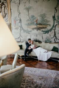 Ein Paar in formeller Kleidung entspannt sich auf einem eleganten Sofa im Vintage-Stil in einem Raum mit einem großen, kunstvollen Wandgemälde. Die Kulisse erinnert an das klassische Ambiente einer traumhaften Hochzeitsplanung, komplett mit Holzböden und sanfter Beleuchtung, die den romantischen Reiz noch verstärkt.