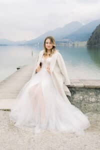 Eine Frau in weißem Kleid und Mantel sitzt auf einem Felsvorsprung neben einem ruhigen See, im Hintergrund sind Berge zu sehen. Der bewölkte Himmel verleiht der Szene eine sanfte, ruhige Atmosphäre – ideal für eine intime Hochzeitsplanung inmitten der Schönheit der Natur.
