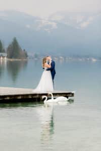 Ein Paar in festlicher Kleidung umarmt sich auf einem Holzsteg an einem ruhigen See, im Hintergrund schneebedeckte Berge. Als Teil ihrer Hochzeit gleiten zwei Schwäne anmutig über das Wasser in der Nähe und verleihen ihrem besonderen Tag einen Hauch von Eleganz.