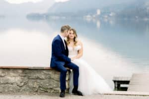 Ein Paar in Hochzeitskleidung sitzt auf einer Steinmauer an einem ruhigen See und fängt die Essenz von „Heiraten“ ein. Der Bräutigam trägt einen marineblauen Anzug, während die Braut ein weißes Kleid trägt. Im Hintergrund ragen neblige Berge auf, während sie sich über die ruhige Wasseroberfläche hinweg liebevoll anschauen.