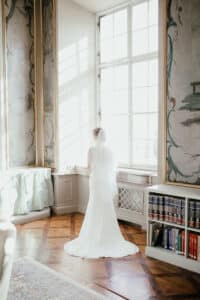 Eine Braut in einem weißen Kleid und Schleier steht in einem sonnendurchfluteten Raum und blickt aus einem großen, verzierten Fenster. Der Raum strahlt mit edlen Holzböden, dekorativen Wandpaneelen und einem Bücherregal Vornehmheit aus und verkörpert die heitere Eleganz, die perfekt für die Hochzeitsplanung ist.