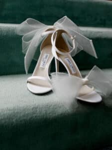 Elegante weiße Sandaletten mit großen Tüllschleifen zieren eine mit grünem Teppich ausgelegte Treppe, perfekt für diese besonderen Hochzeitsmomente.