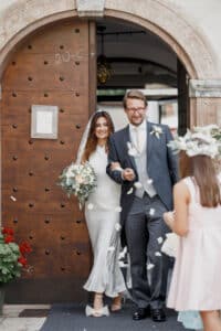 Ein frisch vermähltes Paar kommt durch eine Holztür und lächelt freudig nach seiner Hochzeit. Die Braut hält einen Blumenstrauß und trägt einen Schleier, während der Bräutigam einen Anzug trägt. Ein Kind im Vordergrund wirft Blütenblätter und trägt so zur festlichen Atmosphäre ihrer sorgfältig ausgearbeiteten Hochzeitsplanung bei. Rote Blumen schmücken die linke Seite.